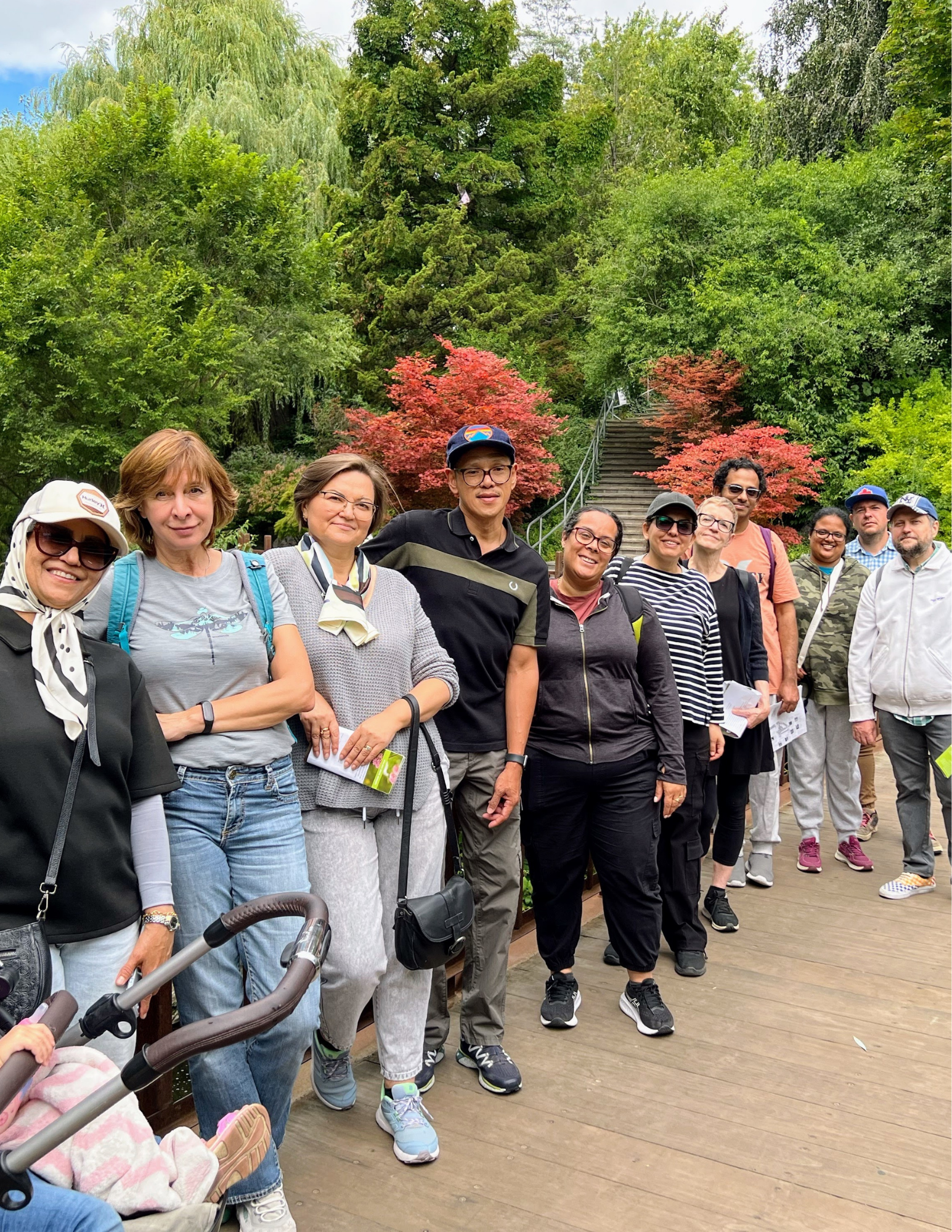 A group of people posing together outdoors on a walkway, with trees and green foliage in the background.