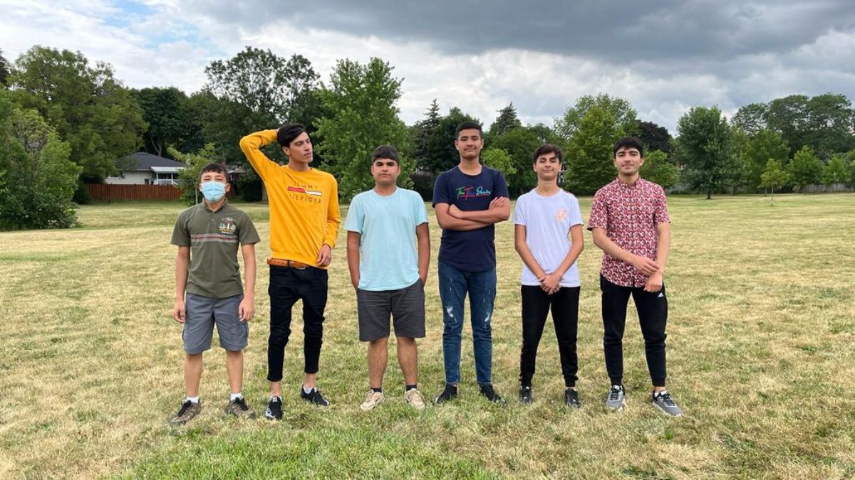 Group of teenagers stand facing the camera. They are in a park. There are trees and cloudy skies behind them.