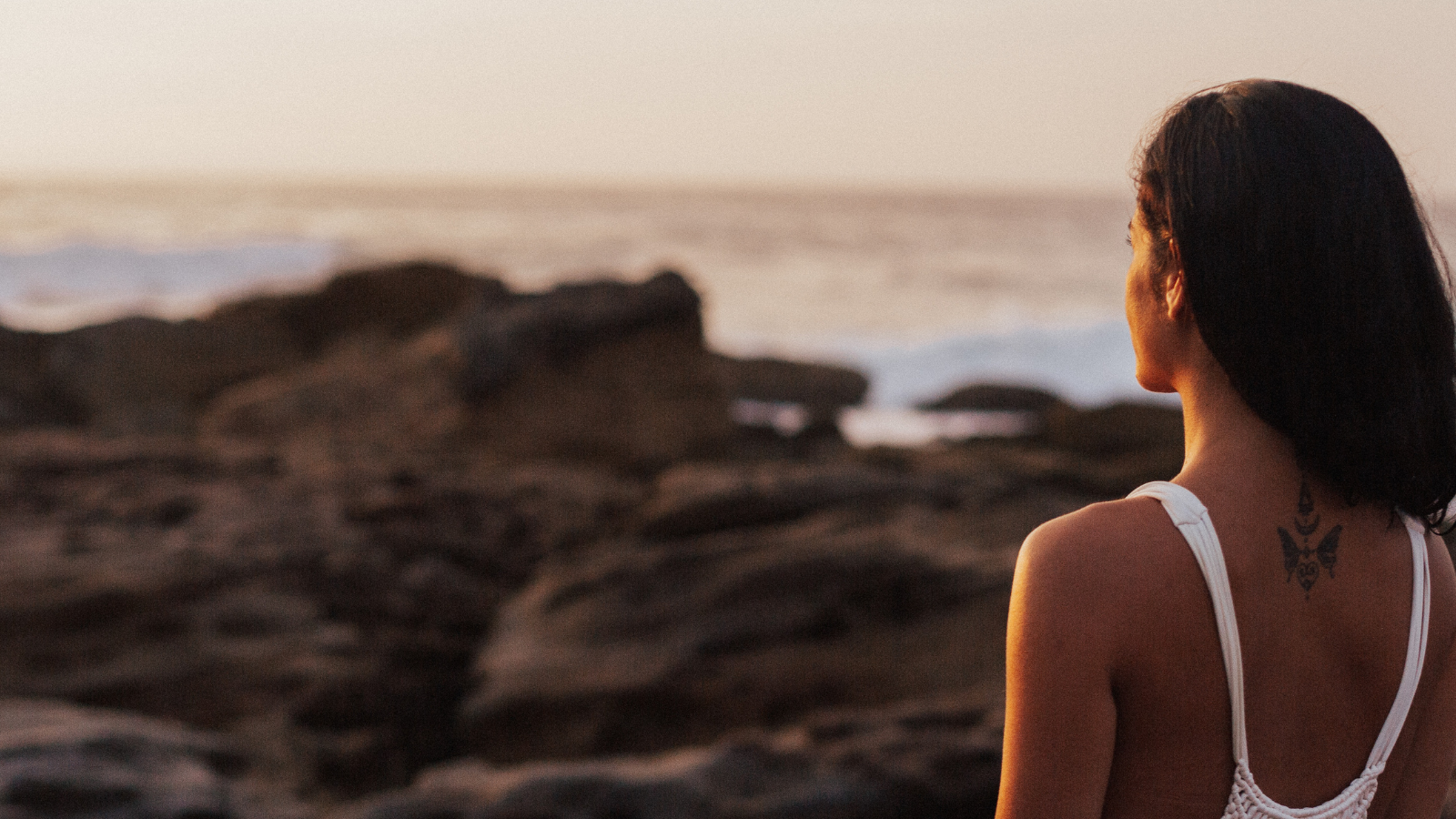 Feminine person stands facing a rocky shore. The photographer captures her back. She has long, dark hair and wears a white tanktop.