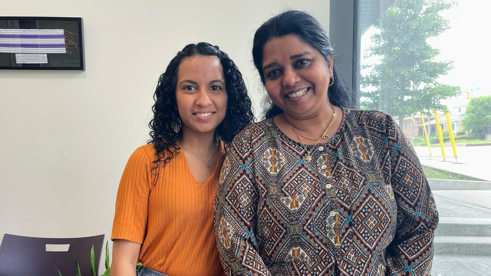 A photograph of placement student Wasana, with mentor Jeya, Community Settlement Lead. Both smile at the camera as natural light comes in through the window.