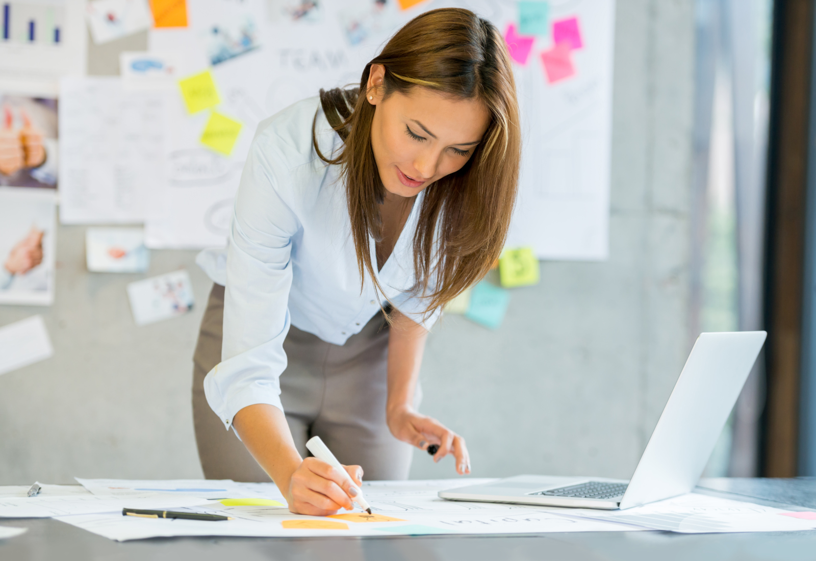 Woman writing on paper in an office with papers and a laptop on the table.
