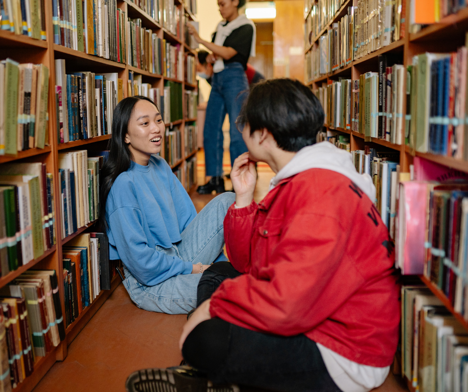 Two people sitting on the floor of a library aisle talking, surrounded by bookshelves, with two others in the background.