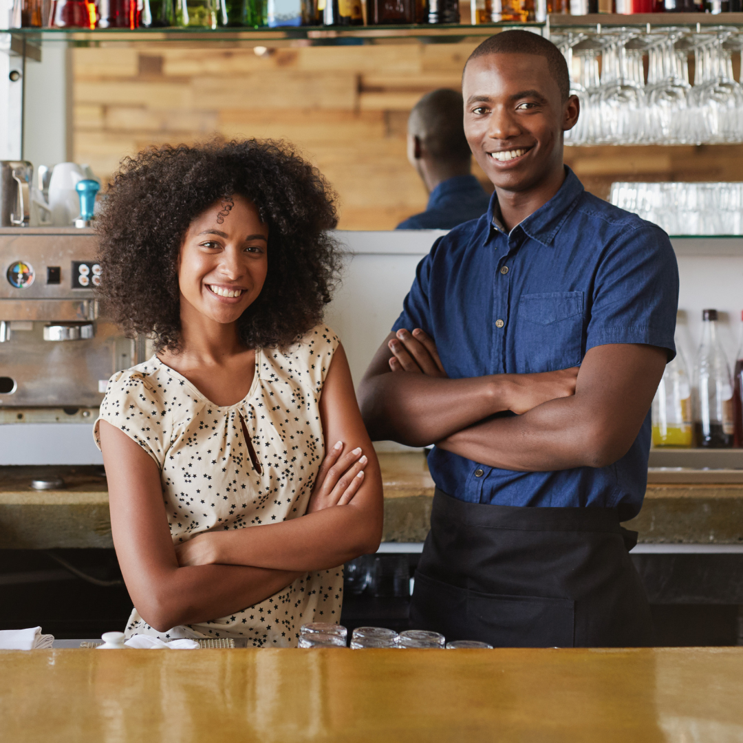 Two people standing behind a cafe counter with arms crossed, smiling.