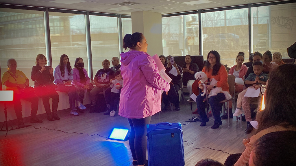 A woman standing in the centre of the room. Beside her is a suitcase. The audience sits in a semi-circle.
