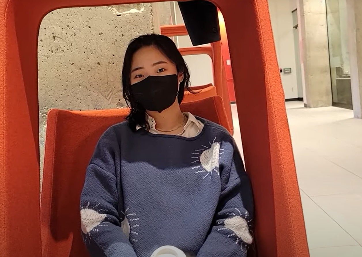 A young person sitting in a red chair facing the camera. They have a face mask on.