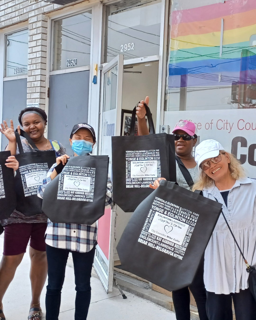 A group of adults standing, smiling and holding up bags that read "Eglinton & Lawrence". They're outside of a city councillors office.
