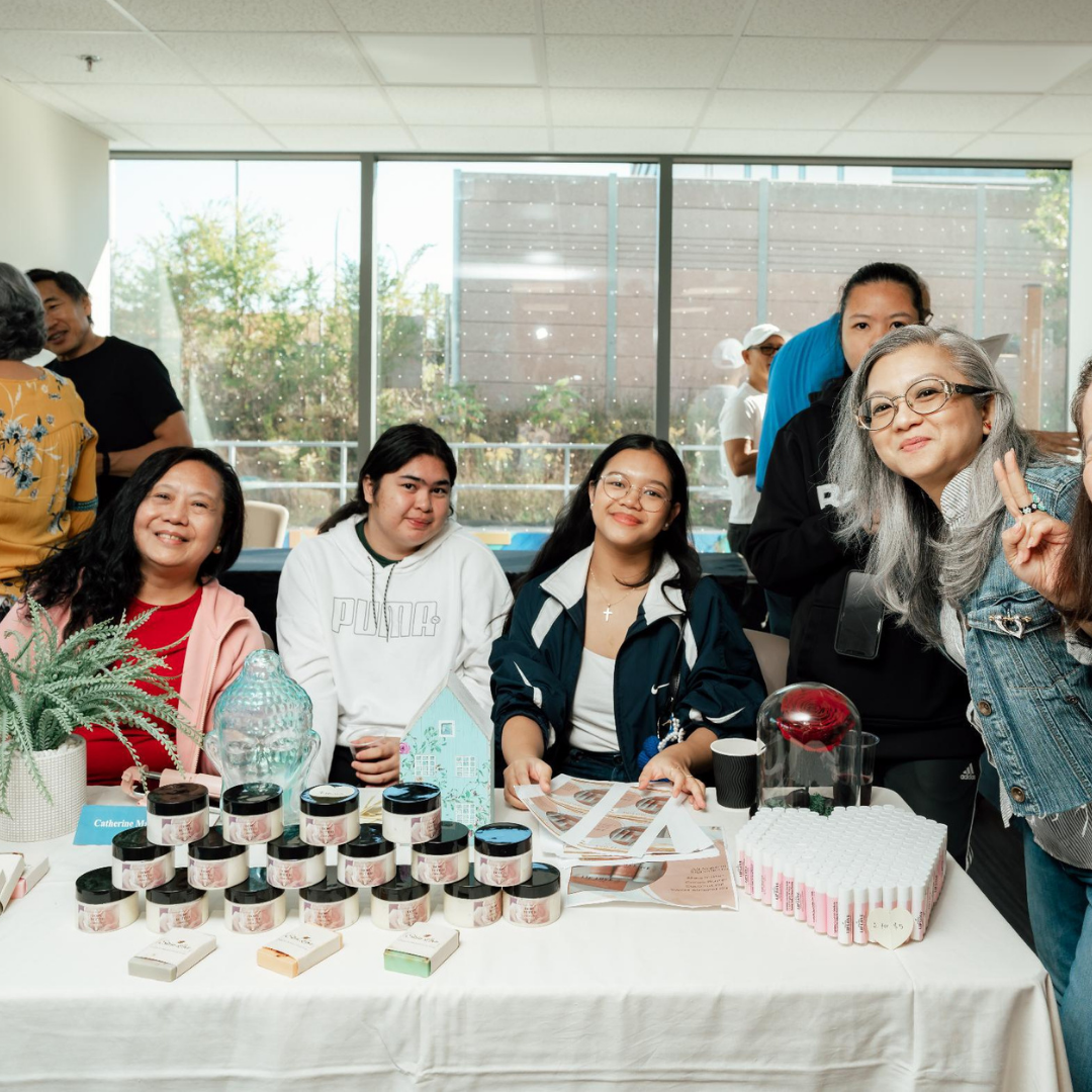 A group of women sit behind a table. On the table is a display of handmade body butter.
