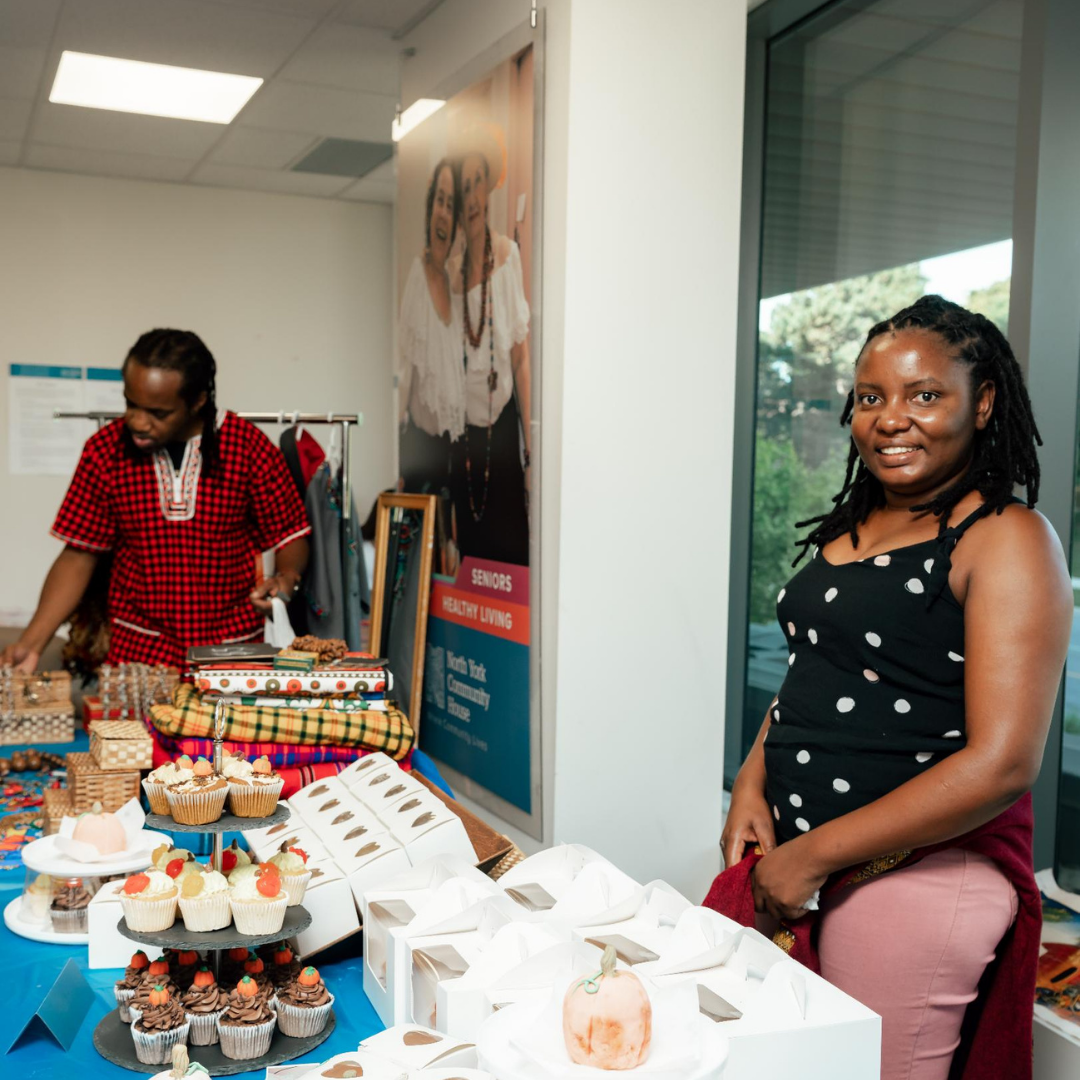 A women smiles at the camera. She stands in front of a table with cupcakes.