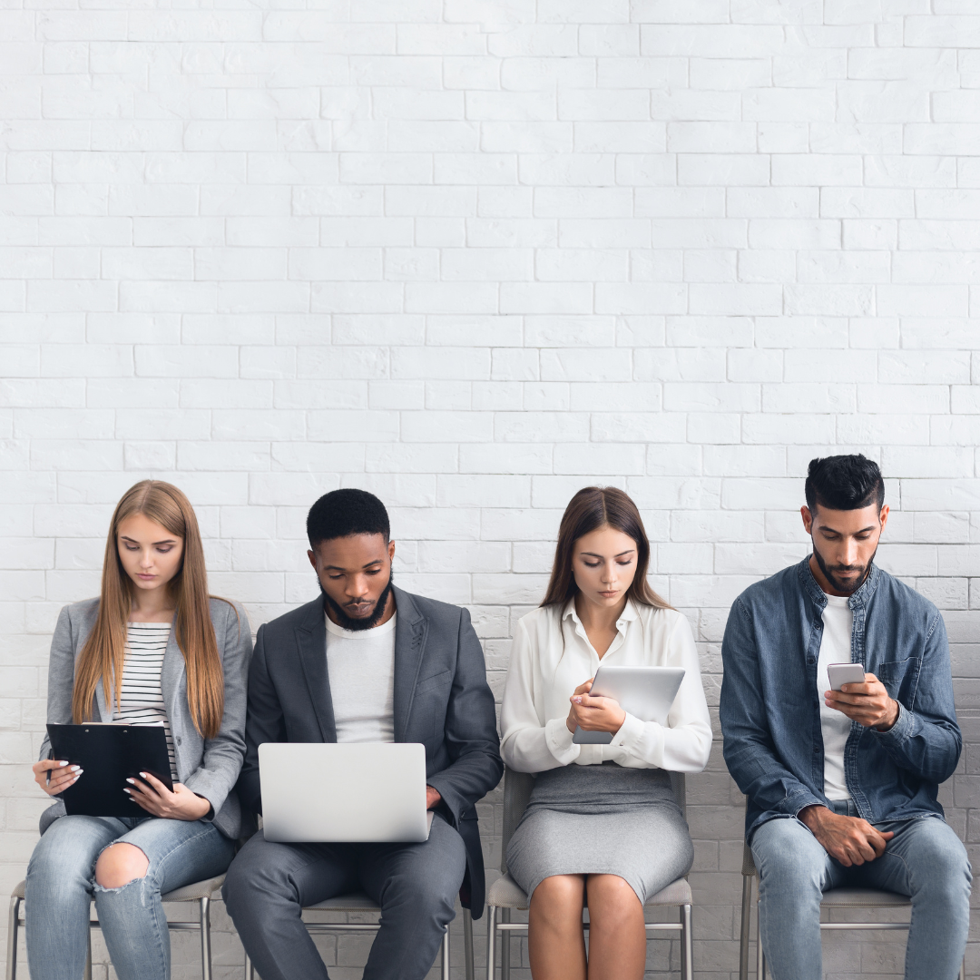 Four people sitting in a row against a white brick wall, using electronic devices.