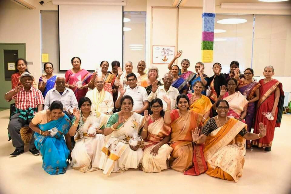 A large group of adults in colorful traditional attire posing indoors, some seated and some standing, smiling and waving at the camera.