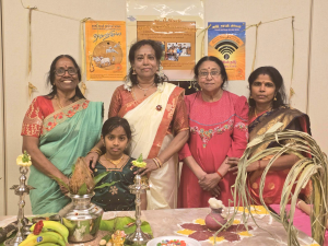 A group of five people in traditional attire standing behind a decorated table, featuring cultural artifacts and Tamil Heritage Month posters in the background.
