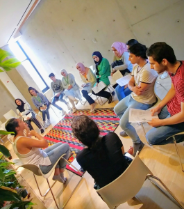 A group of people sitting in a circle in a modern room having a discussion with a geometric-patterned rug.