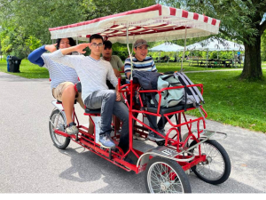 Four youth riding a red quadracycle with a striped canopy on a park path.