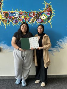 Adriana and Farwa holding a certificate in front of a colorful mural.