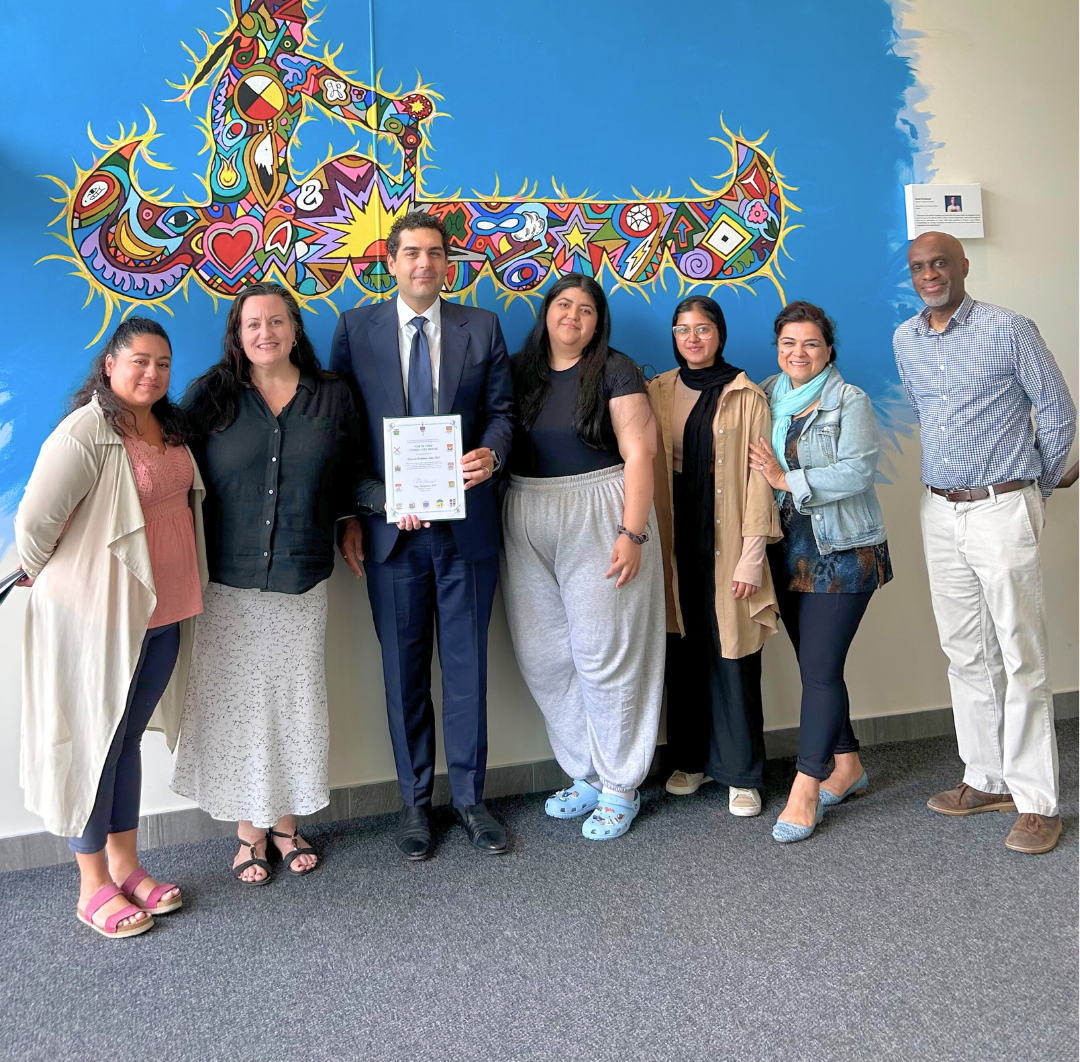 A group of seven people stands in front of a colorful abstract mural, with one person holding a certificate.