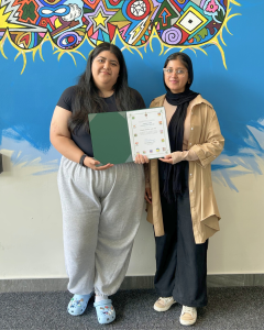 Adriana and Farwa holding a certificate in front of a colorful mural.