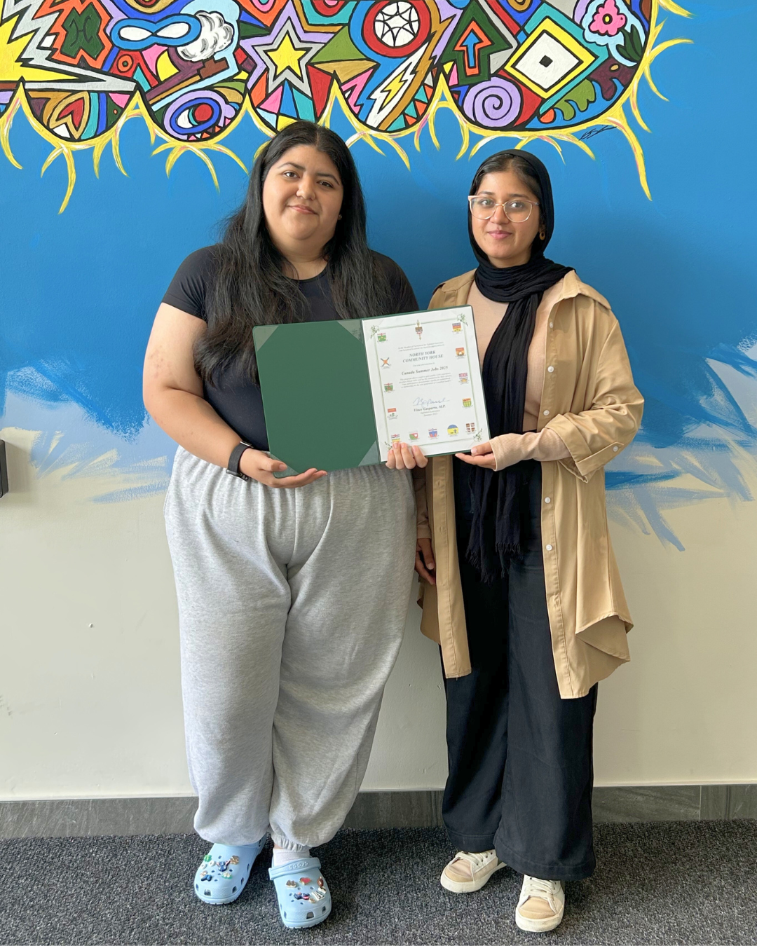 Adriana and Farwa holding a certificate in front of a colorful mural.