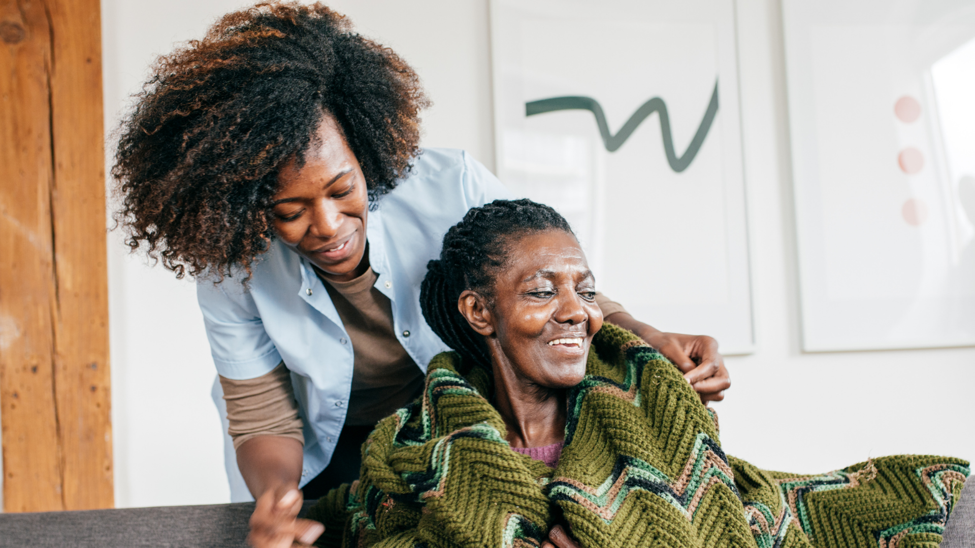 A younger person helps an elderly person with a green knitted blanket in a cozy room.