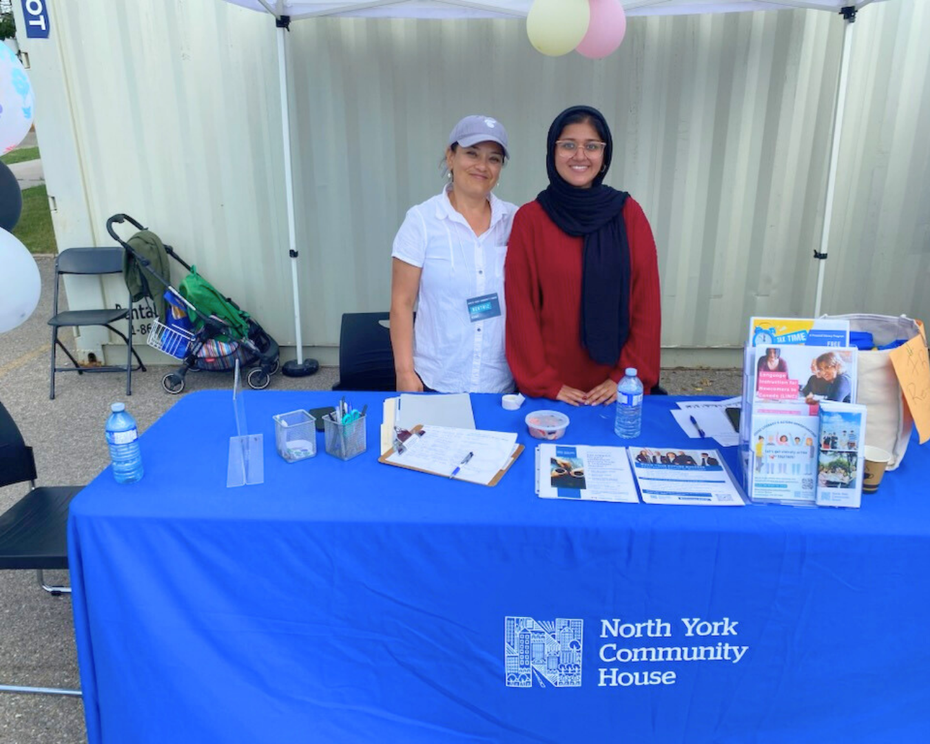 Beatriz and Farwa at an information booth with a blue North York Community House tablecloth and various materials on the table.