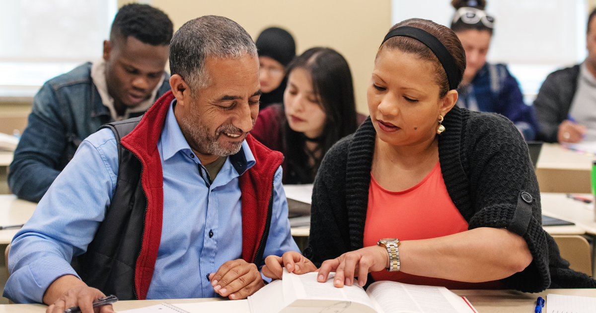 Man and woman discussing a book in a classroom with others in the background.