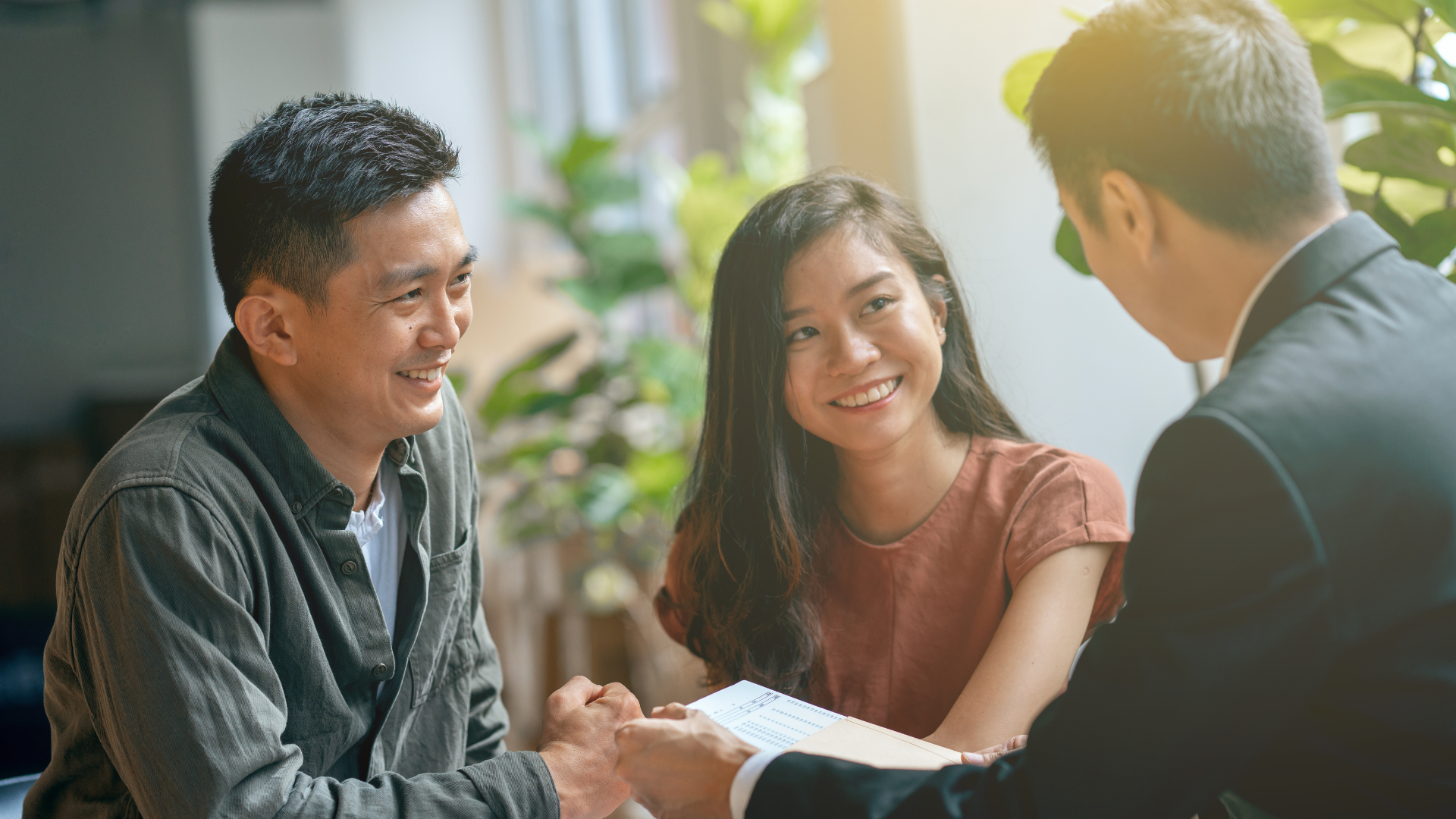 Three people having a friendly conversation at a table, surrounded by plants and natural light.
