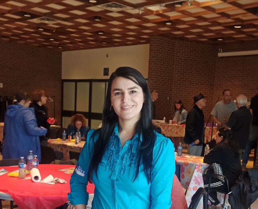 Zahra in turquoise shirt smiling indoors at a social event with people gathered around tables.