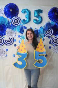 Zahra smiling, holding large cutouts of the number 35 with party hats in front of a blue and white decorated backdrop.