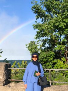 A picture of Aliya standing outside, in front of a tree and a rainbow.