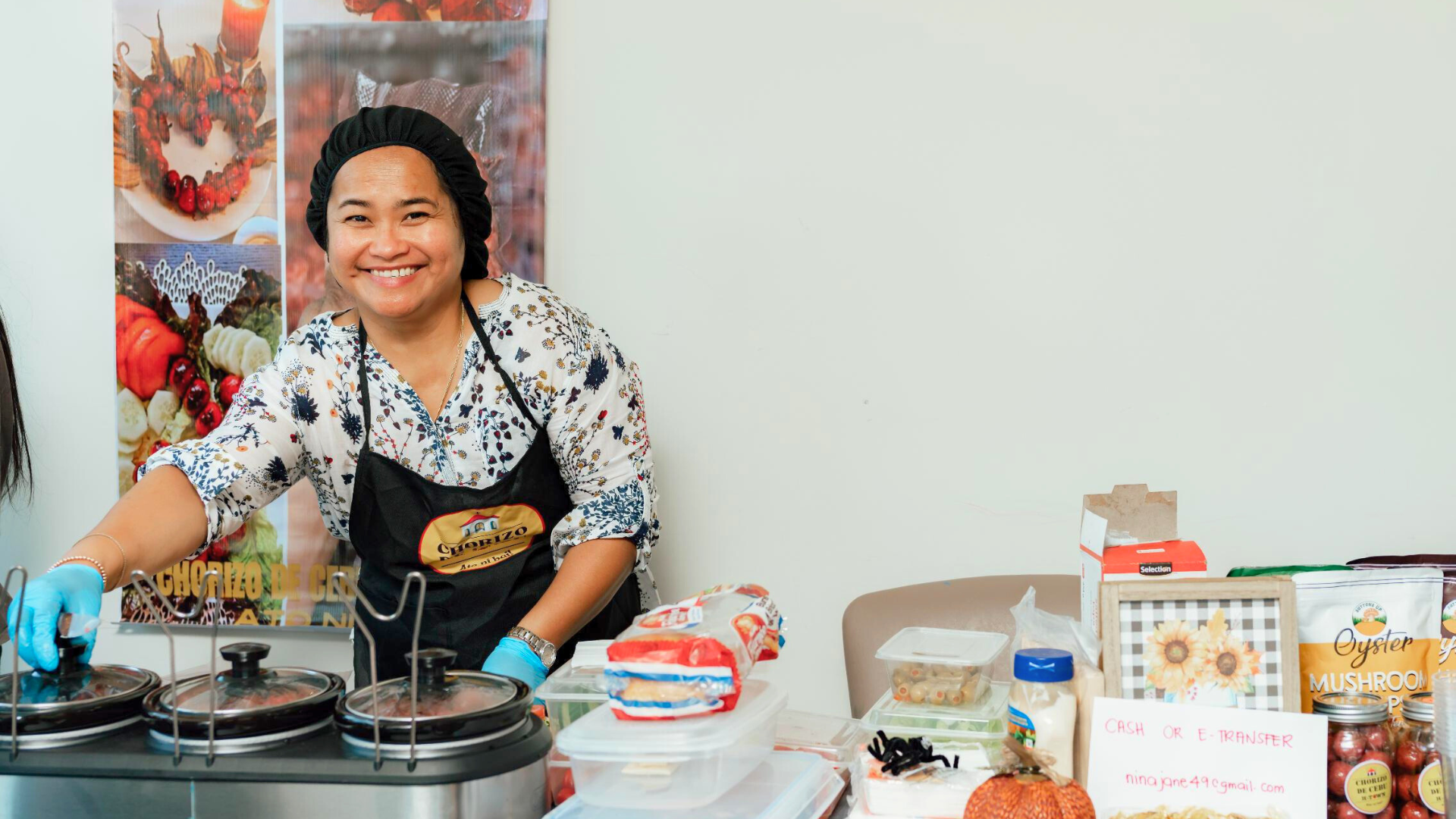 A woman in a floral blouse and apron stands behind a table with food items and slow cookers.