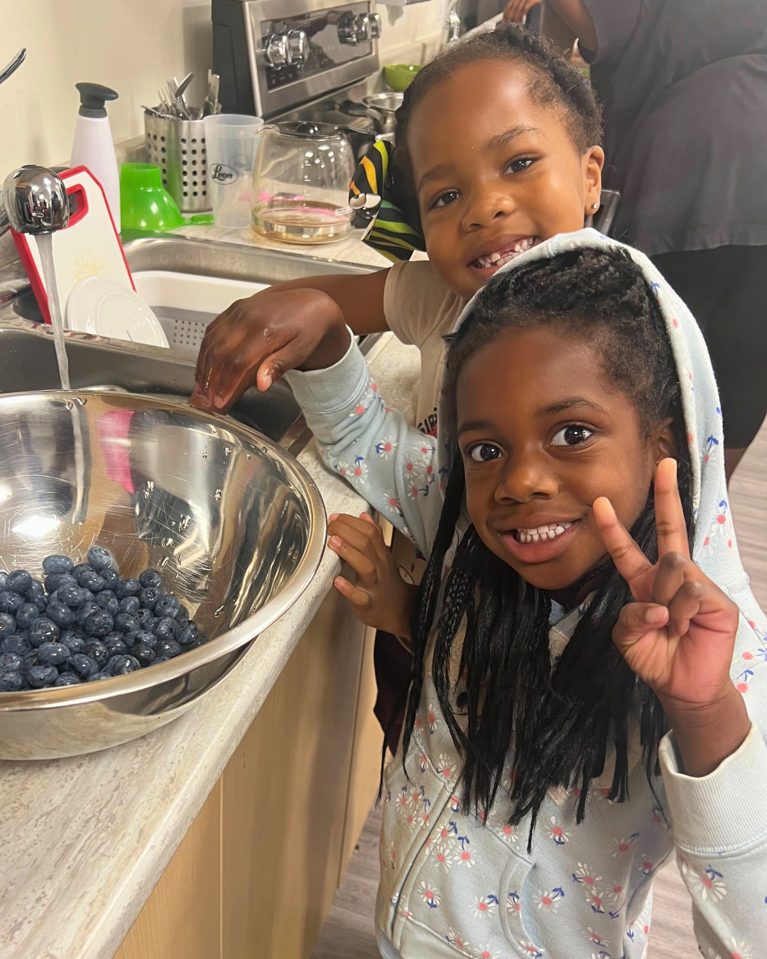 Two children smiling by a kitchen counter with a bowl of blueberries.