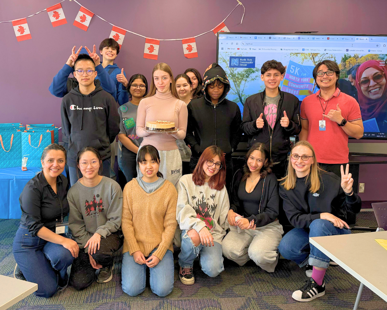 A diverse group of people pose smiling in a room with purple walls and Canadian flag bunting, some holding a cake and making peace signs.