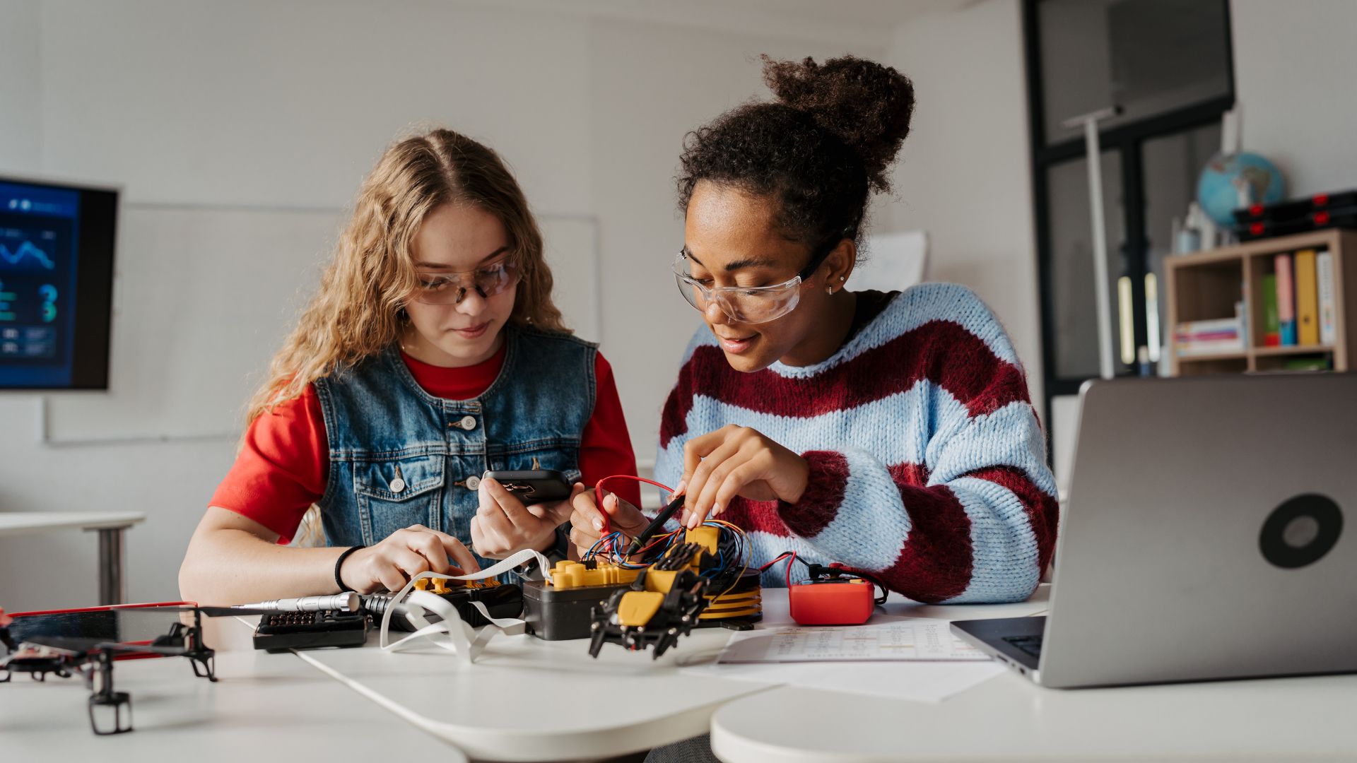 Two youth in safety glasses work on electronics at a table, with a laptop and a small drone nearby in a classroom.