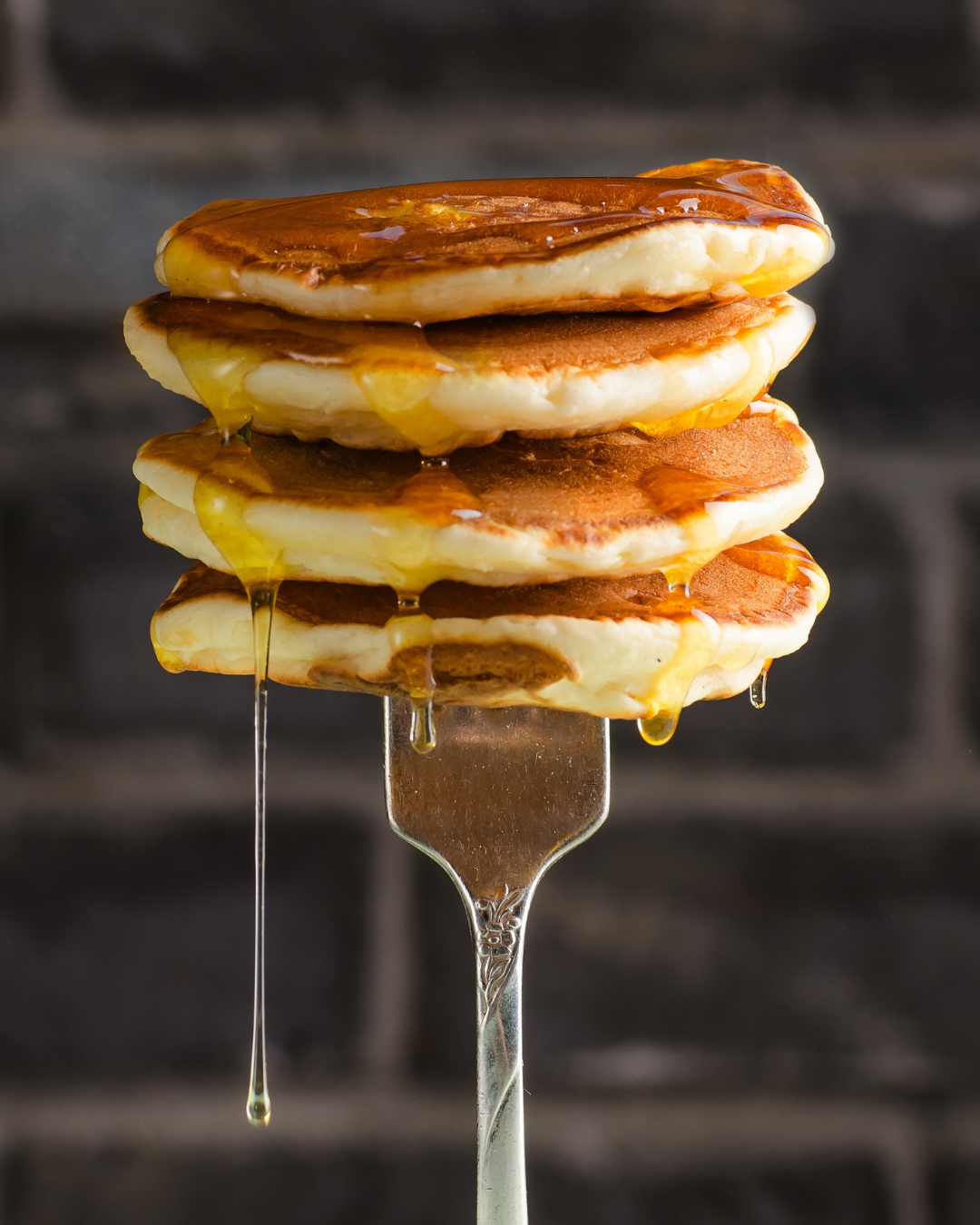 A stack of pancakes drizzled with syrup on a fork against a brick wall background.