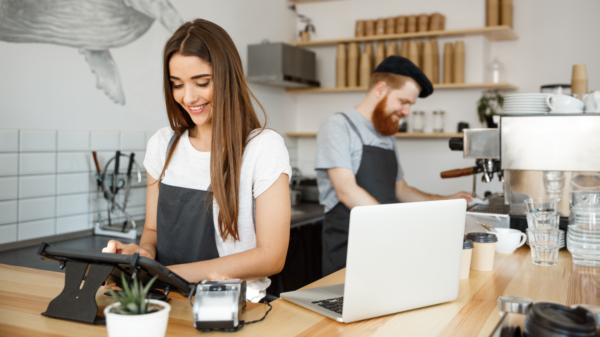 Baristas working in a coffee shop with a wooden counter, a laptop, and an espresso machine.