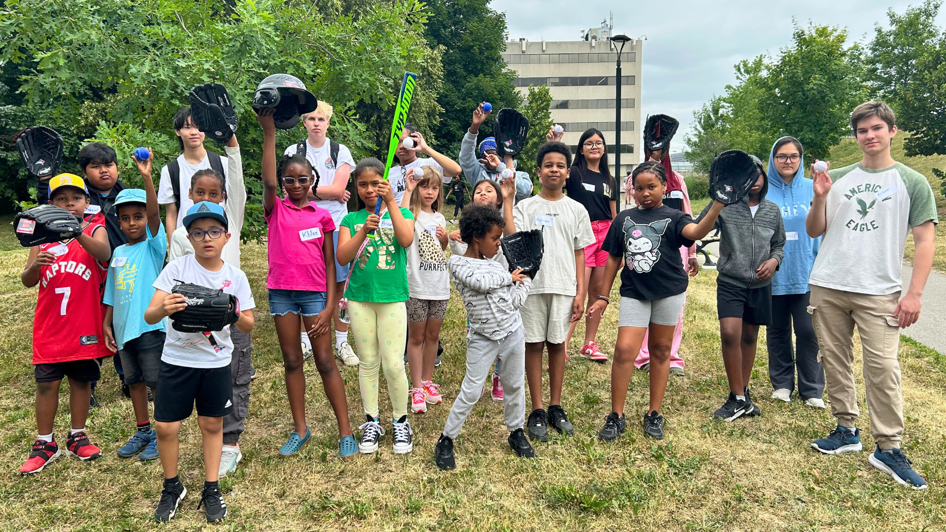 A group of children and adults pose with baseball equipment in a grassy area.