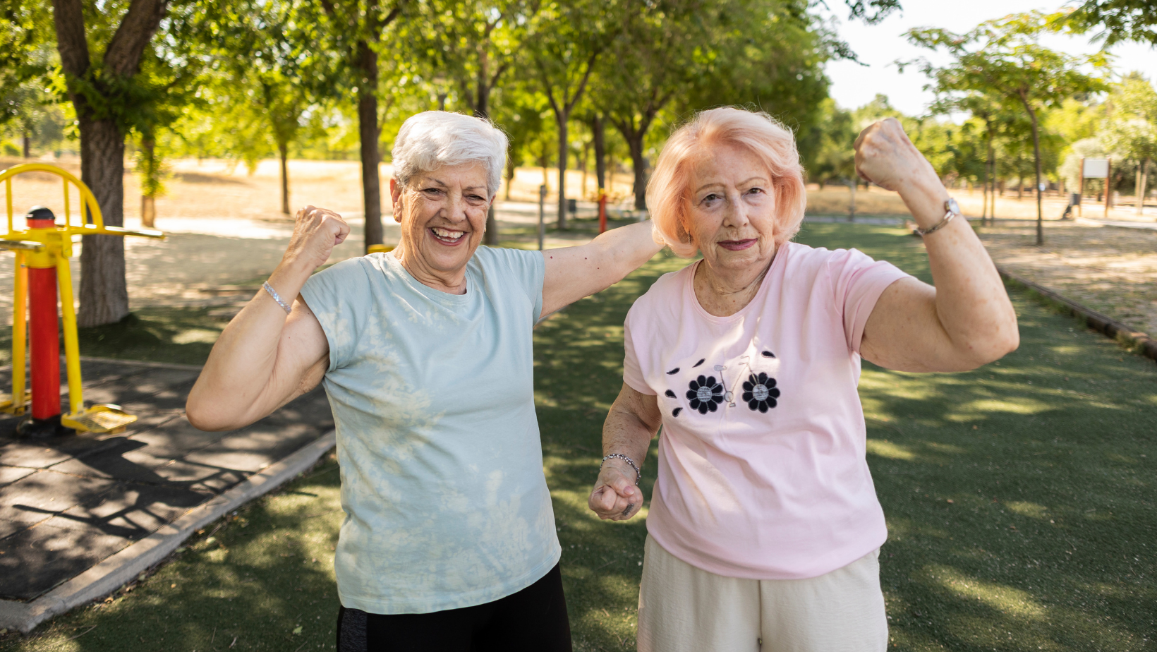 Two older adults smiling and flexing their arms in an outdoor park.