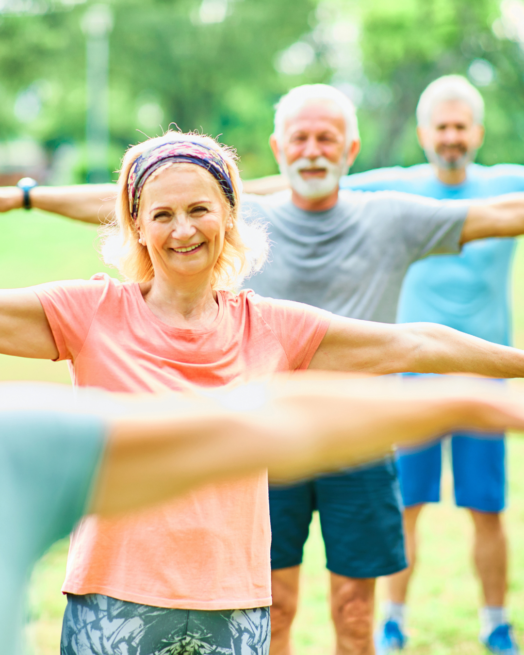 Older adults exercising outdoors with arms outstretched, focusing on a smiling woman in a peach T-shirt.