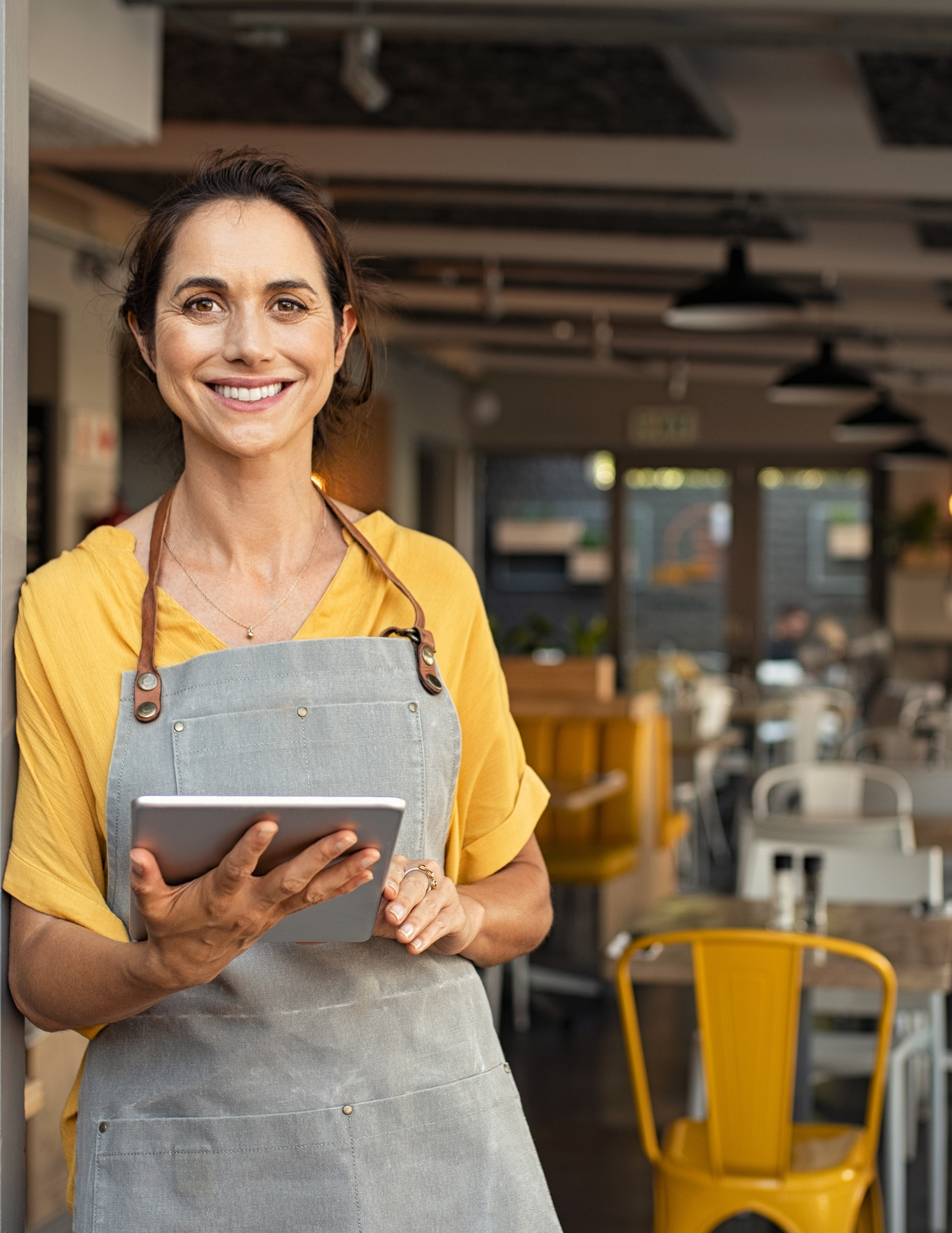 A woman stands in the doorway of a restaurant holding an ipad and smiling.