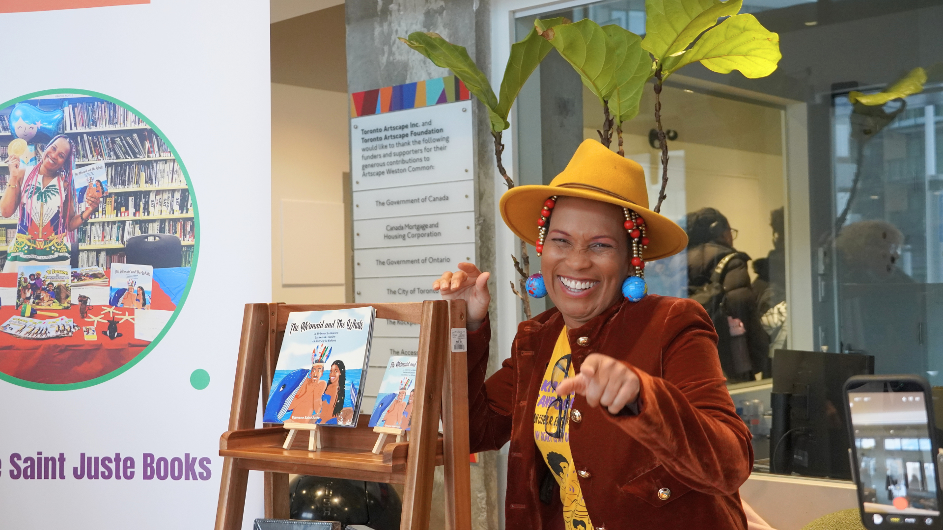 A woman smiles and points to the camera. She is standing next to a shelf with a book on it.