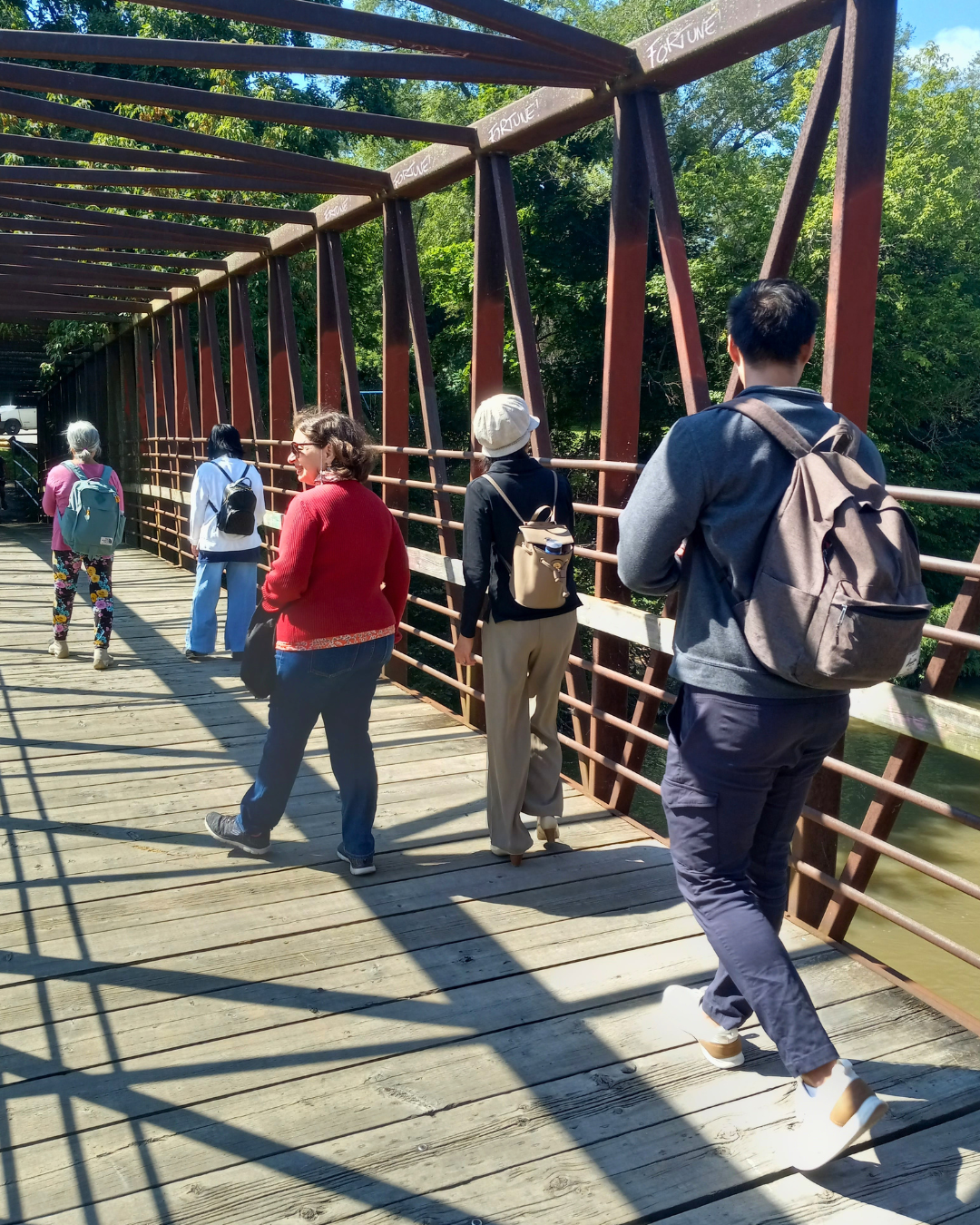 People walking on a wooden footbridge with metal beams and railings, surrounded by green trees.
