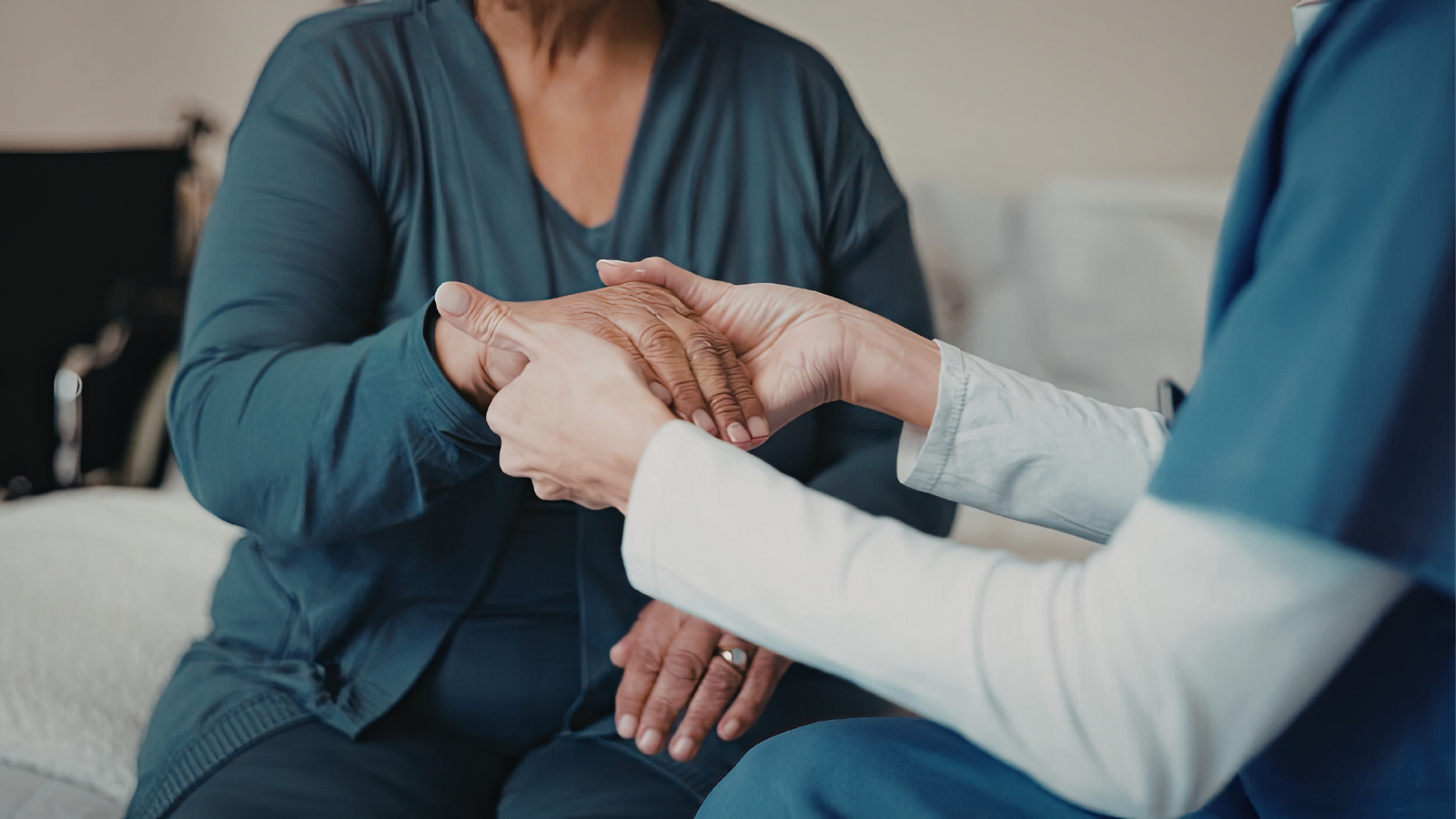 A caregiver in a blue uniform holds the hands of an elder sitting on a bed.