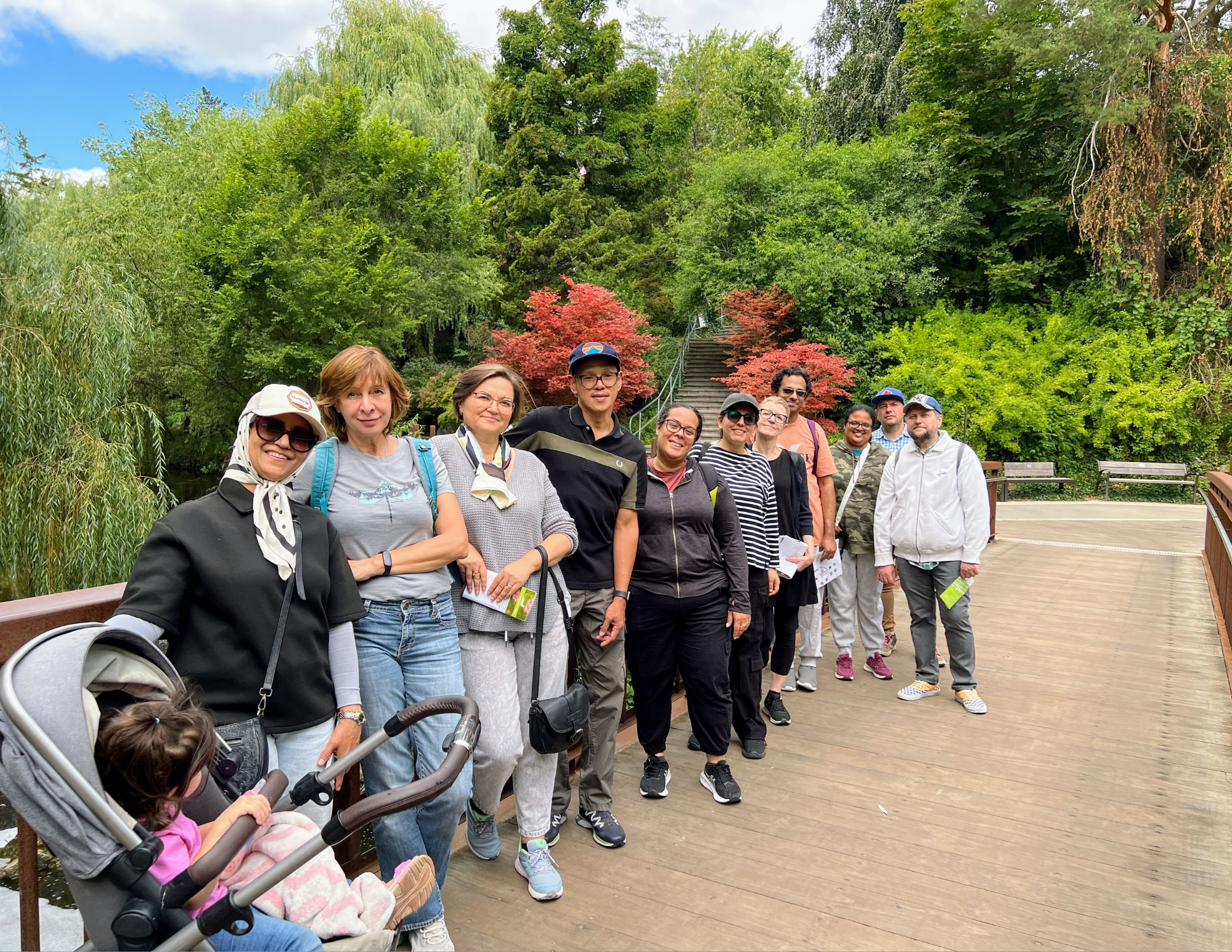 A group of people posing together outdoors on a walkway, with trees and green foliage in the background.