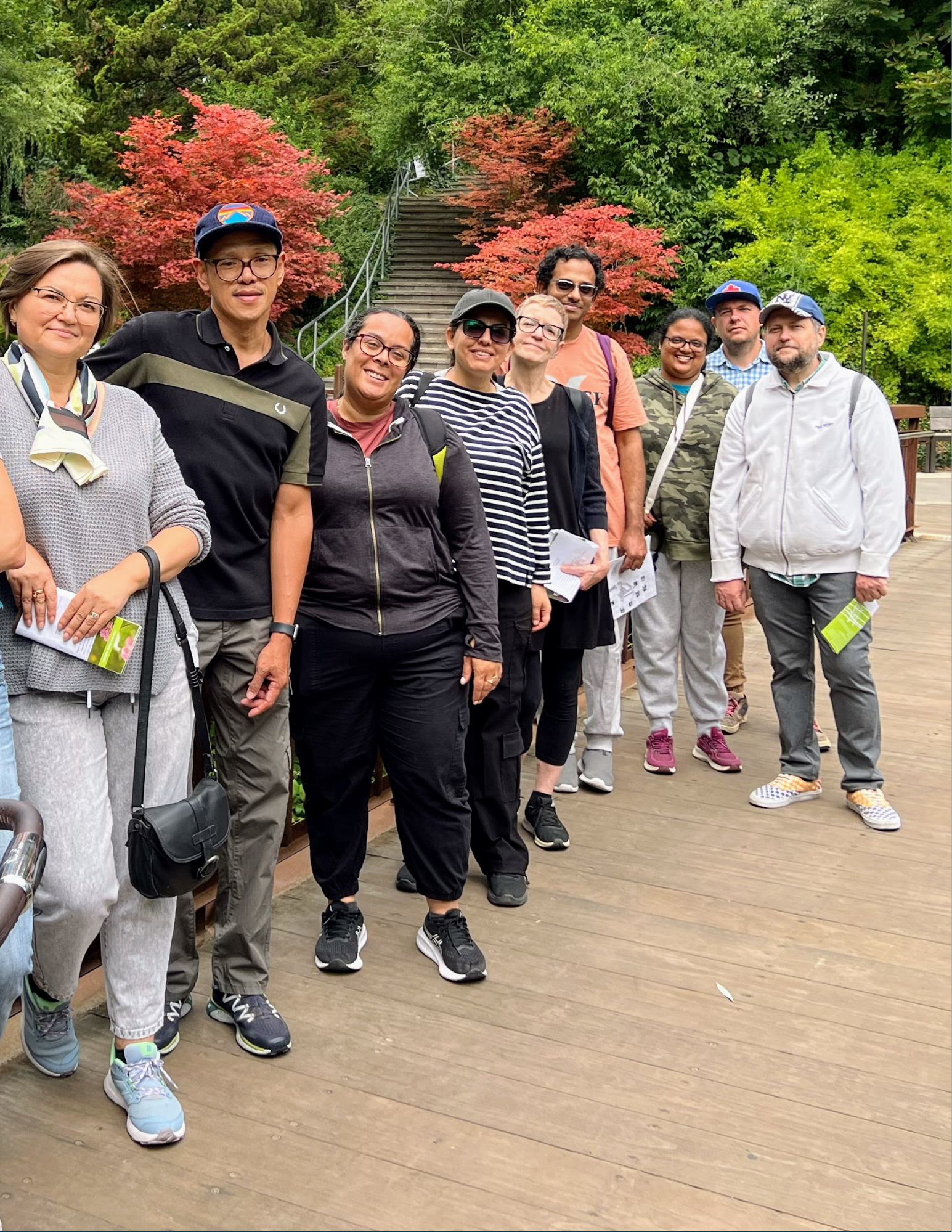 A group of seven people posing together outdoors on a walkway, with trees and green foliage in the background.