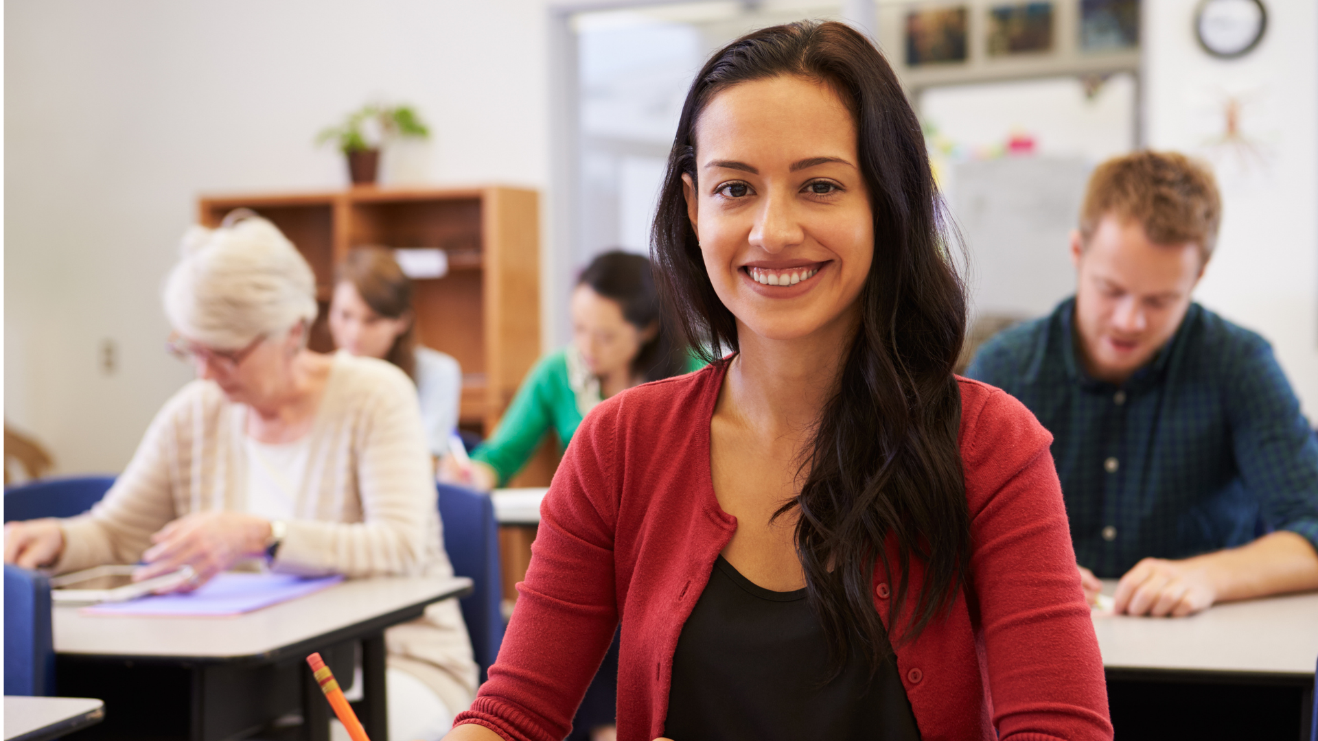 Smiling woman in a red cardigan seated in a classroom with other people working at desks behind her.