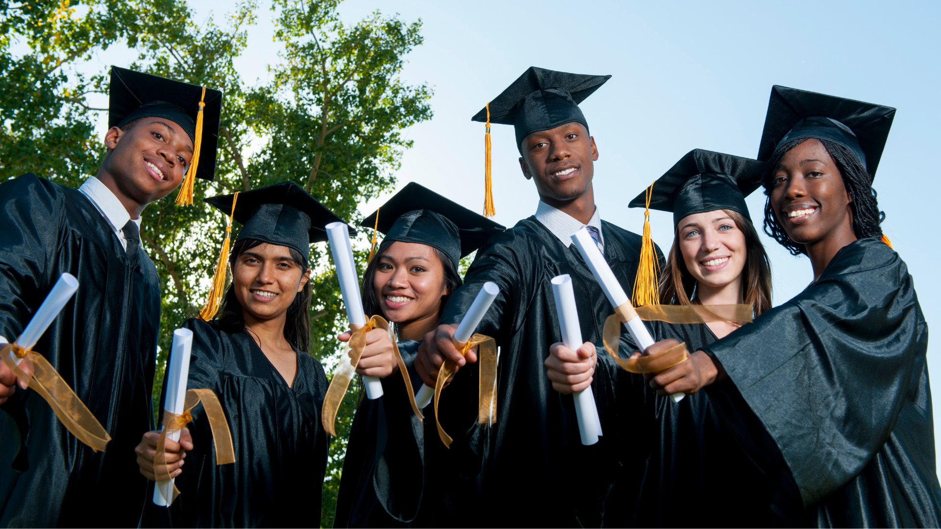 Six graduates in black caps and gowns hold rolled diplomas outdoors.