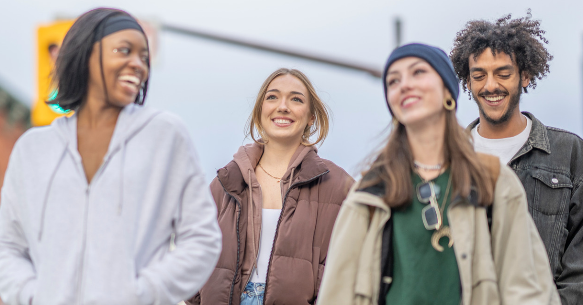 Four smiling young adults stand together outdoors, wearing casual clothing.